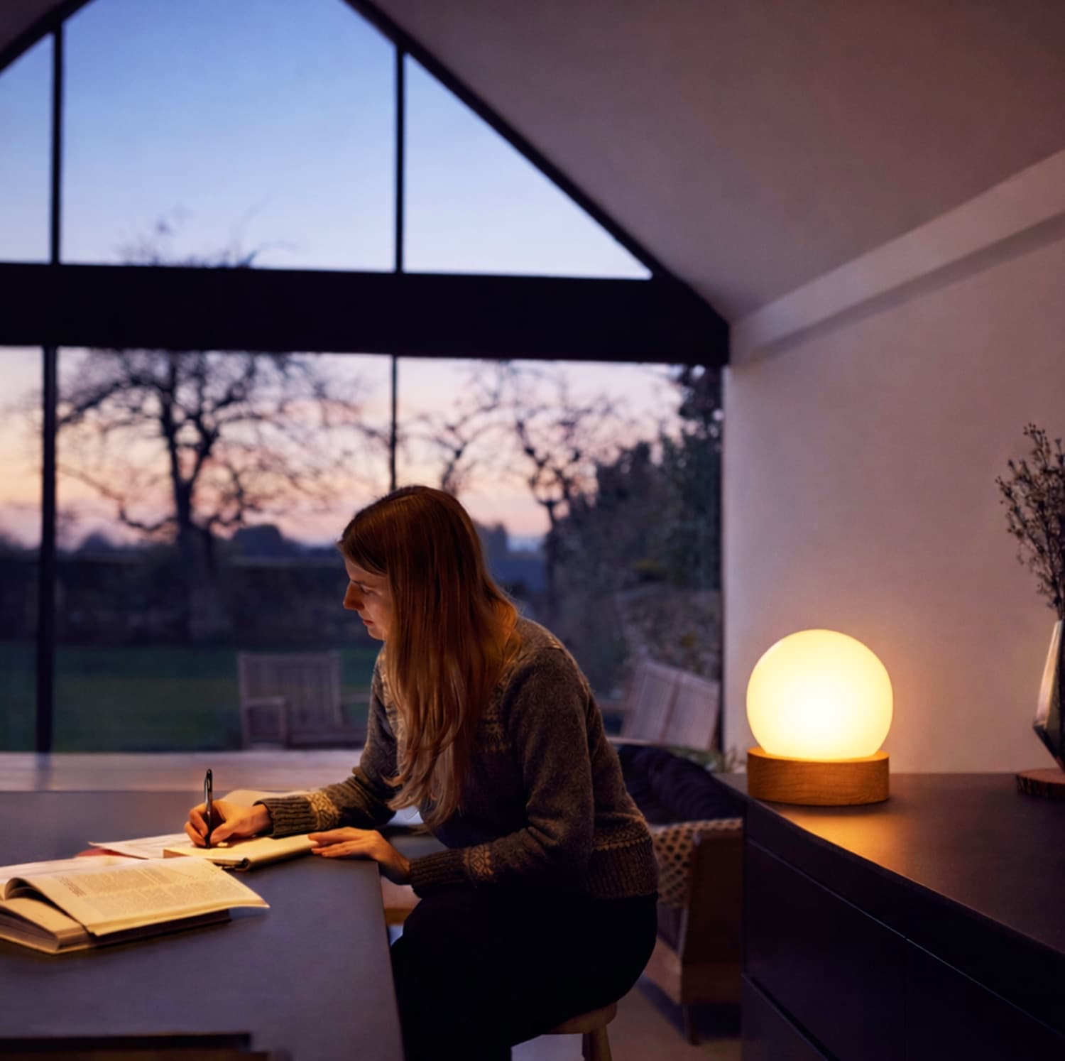 An OIO lamp glowing warm and blue-free at dusk beside someone reading, sunset fading in the window behind.